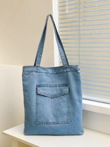 This is a light blue denim tote bag with a small front pocket and two long straps. It’s kept on a white table near a window with blinds.